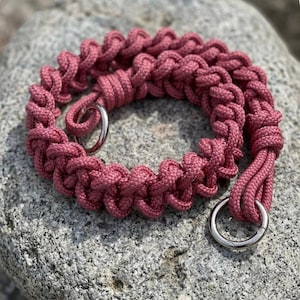 Könnte beinhalten: Ein altrosa Paracord-Halsband mit einem dicken, geflochtenen Muster. Das Halsband verfügt über zwei polierte, silberfarbene Metallringe an den Enden sowie ein sicheres Knotendetail, das die geflochtenen Kordelabschnitte miteinander verbindet.