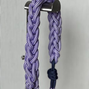 以下が含まれることがあります： 紫と白の濃淡の編みロープのハルター、ネイビーブルーのアクセント付き。 ハルターは、クラシックな編み模様で設計されており、金属製のバーから吊り下げられています。 ロープは太く、耐久性があるように見えます。