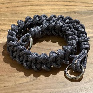 May include: A gray braided rope camera strap with two silver metal rings.