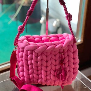 Op de afbeelding: Een felroze gebreide handtas met een grove breistructuur. De tas heeft een lange roze band en een vredesduif-bedel.