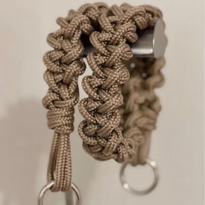 May include: A tan paracord strap featuring a complex, braided weave pattern. The strap is constructed from durable, woven nylon cord and includes two circular silver-toned metal rings attached at the ends, providing a secure connection point.