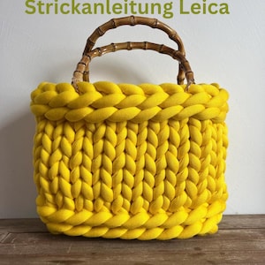 Könnte beinhalten: Eine leuchtend gelbe, handgestrickte Tragetasche mit Bambusgriff. Die Tasche hat ein dickes, geflochtenes Design an den oberen und unteren Kanten, und der Text "Strickanleitung Leica" ist oben sichtbar. Die Tasche ist aus weichem, dickem Garn gefertigt.