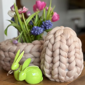 Könnte beinhalten: Ein hellbrauner gestrickter Korb gefüllt mit einem Strauß rosa und lila Blumen. Eine grüne Keramik-Hasenfigur steht vor dem Korb.