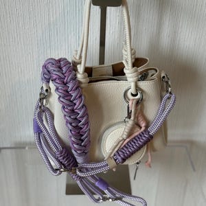 Könnte beinhalten: Cremefarbene Handtasche mit geflochtenem lila-weißem Riemen. Die Tasche hat eine kleine, runde Reißverschlusstasche und ein passendes geflochtenes Handgelenkband. Silberfarbene Hardware akzentuiert die Tasche und den Riemen.