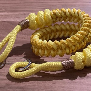 Könnte beinhalten: Ein gelber geflochtener Schlüsselband mit einem silbernen Ring und einem goldenen Akzent. Das Seil ist in einem komplexen Muster geflochten.