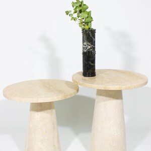 May include: Two beige stone side tables with round tops and conical bases. One table has a black and white marble vase with green foliage on top. The tables are made of a light-colored stone.