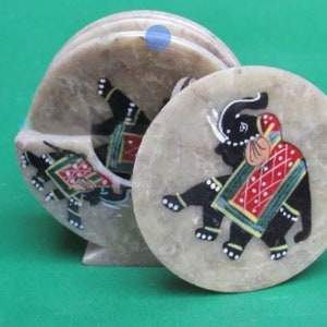 May include: A set of beige stone coasters with an elephant design. The elephant is black with red, green, and yellow accents. The coasters are round and come with a holder.