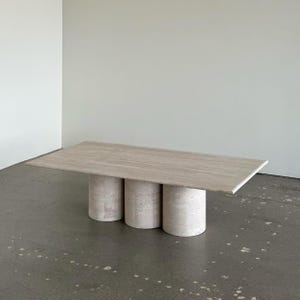 Op de afbeelding: Een rechthoekige tafel van lichtgekleurde steen met een glad oppervlak. De tafel wordt ondersteund door drie cilindrische stenen basissen. Het ontwerp is minimalistisch en modern, geschikt voor een woon- of eetkamer.