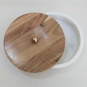 Puede incluir: Un recipiente decorativo redondo con tapa de madera e interior blanco. La tapa es de madera marrón claro con un pomo dorado. El recipiente está parcialmente abierto, revelando el interior blanco.
