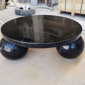Puede incluir: Una mesa de centro redonda de mármol negro con vetas blancas. La encimera descansa sobre dos bases esféricas. La mesa está hecha de piedra pulida y es adecuada para uso en interiores. El diámetro de la mesa es de aproximadamente 122 cm.