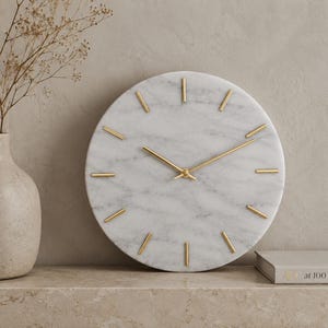 Puede incluir: Un reloj redondo de mármol con manecillas y marcadores dorados. La esfera del reloj es blanca con vetas grises. El reloj está sobre una superficie de color neutro.