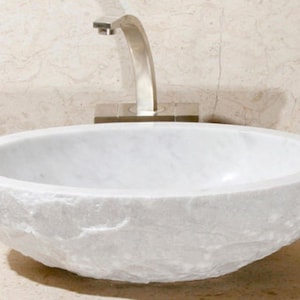 Peut inclure: Un lavabo de salle de bain en marbre blanc avec un extérieur rugueux et texturé. Le lavabo est de forme ovale et a un intérieur lisse et blanc.