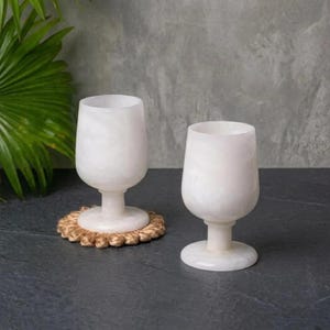 May include: Two white, translucent wine glasses with a stem and round base. One glass rests on a round, woven coaster. The glasses are set against a dark surface with a gray background and a green plant.
