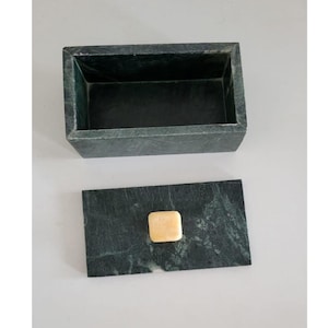 Può includere: Una scatola rettangolare in marmo verde scuro con coperchio abbinato. Il coperchio presenta un piccolo pomello quadrato di colore chiaro. La scatola è aperta, rivelando un interno scuro. Il marmo ha venature naturali.