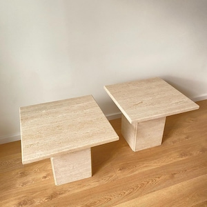 Puede incluir: Dos mesas auxiliares cuadradas de piedra travertina de color beige claro. Las mesas tienen un diseño sencillo con una tapa cuadrada y una base cuadrada.
