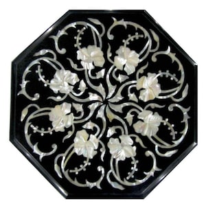 May include: An octagonal black table top with a floral design inlaid with mother-of-pearl. The design features white flowers and swirling vines radiating from the center, creating a symmetrical pattern. The edges are smooth and polished.