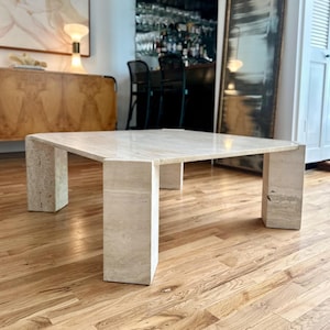 Puede incluir: Una mesa de centro cuadrada de travertino en color beige claro. La mesa tiene un diseño geométrico sólido con cuatro patas rectangulares. Está colocada sobre un suelo de parquet. Dimensiones aproximadas: 100 cm x 100 cm.
