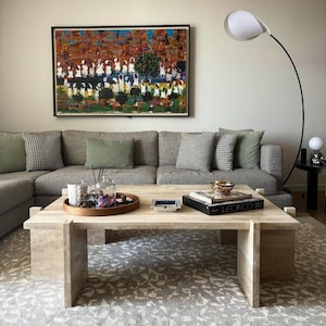 May include: A travertine coffee table with a rectangular top and a unique design. The table has a wooden tray on top with decorative items, including candles and a glass vase. A book titled "AB CONCEPT" is on the table, along with a gold bowl. The table is in a living room setting with a grey sectional sofa and a white floor lamp.