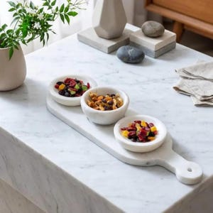 Puede incluir: Una bandeja de mármol blanco con tres cuencos pequeños y redondos llenos de frutos secos y nueces. La bandeja tiene forma rectangular con un asa. Los cuencos están dispuestos en fila, ideal para servir aperitivos.