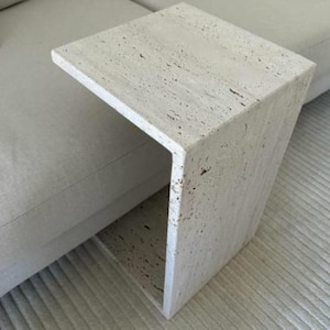 Puede incluir: Una mesa auxiliar de piedra travertina blanca con un diseño único y abierto. La mesa tiene una tapa rectangular y una base abierta y curvada.
