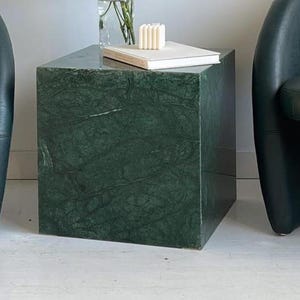 Può includere: Un tavolino quadrato in marmo verde scuro con un motivo venato naturale. Un libro bianco, una piccola pila di candele bianche e un vaso trasparente con fiori sono appoggiati sopra. Il tavolo è accanto a una poltrona verde scuro.