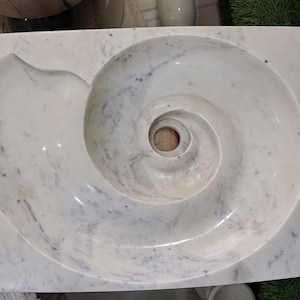 May include: A white marble sink with a nautilus shell design. The sink has a rectangular shape with a spiral basin and a smooth, polished surface. Gray veining adds detail to the white marble.