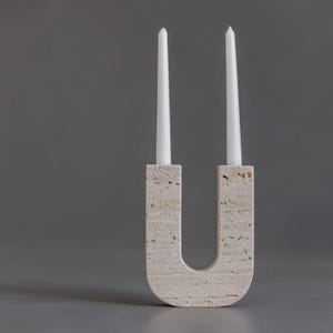 Puede incluir: Un portavelas beige en forma de U hecho de piedra texturizada. Dos velas cónicas blancas están insertadas en el soporte. El portavelas es un elemento decorativo para el hogar.
