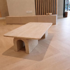 Puede incluir: Una mesa de centro rectangular beige con esquinas redondeadas. La mesa está sostenida por dos bases arqueadas. La mesa está hecha de un material de piedra de color claro, y el diseño es minimalista y moderno. La mesa es un punto focal en un salón.