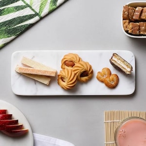 Könnte beinhalten: Ein rechteckiges Serviertablett aus Marmor mit einer Auswahl an Keksen und Gebäck. Das Tablett enthält Mandelgebäck, herzförmige Palmiers, Waffelgebäck und einen Sandwichkeks. Eine kleine Schüssel mit Keksen befindet sich in der oberen rechten Ecke.