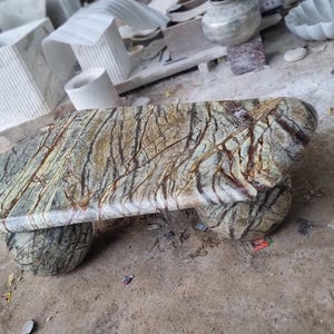 Op de afbeelding: Een stenen bank met een natuurlijk, aards kleurenpalet. De bank heeft een rechthoekige bovenkant met afgeronde randen, ondersteund door twee afgeronde bases. Het oppervlak heeft een uniek patroon van bruine en groene adering.