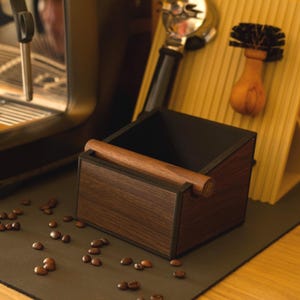 Op de afbeelding: Een houten espresso knock box met een donker interieur en een houten bar. De doos staat op een donkere ondergrond met verspreide koffiebonen. Een koffiemachine en een borstel staan op de achtergrond.