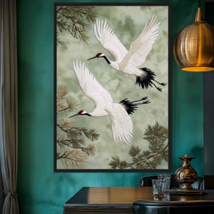 Puede incluir: Obra de arte enmarcada con dos grullas blancas en vuelo sobre un fondo verde y gris suave. Las grullas tienen marcas rojas en la cabeza y plumas negras en la cola. Se ven ramas de pinos.