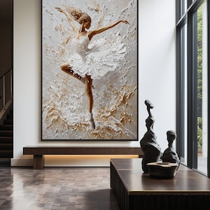 May include: A framed painting of a ballerina in a white tutu, dancing against a textured background of white and gold paint. The painting is hanging on a white wall in a modern interior.