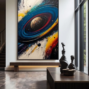 May include: Abstract painting of a colorful galaxy with a planet in the center. The painting is framed in black and hanging on a white wall in a modern interior.