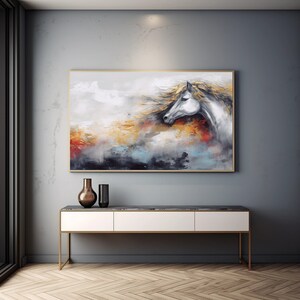 May include: Abstract painting of a white horse's head with a flowing mane, set against a background of muted colours, including shades of grey, blue, and gold. The painting is framed in a simple, gold frame and hangs on a grey wall above a white console table.