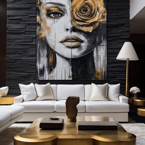 May include: A white sofa with gold throw pillows sits in front of a black wall with a large abstract painting of a woman's face with a gold rose covering one eye. A gold coffee table with a unique shape sits in front of the sofa.