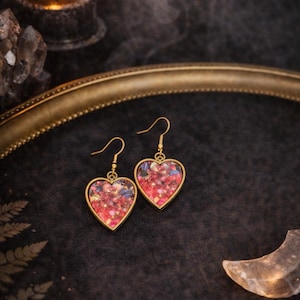 Könnte beinhalten: Herzförmige Ohrringe mit goldfarbenen Rahmen. Die Ohrringe sind mit roten, rosa und blauen floralen Elementen gefüllt. Im Hintergrund befinden sich eine brennende Kerze, ein dekoratives Tablett und Kristalle.