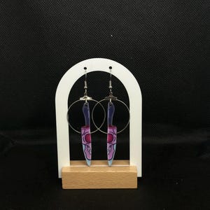 Può includere: Orecchini a cerchio argentati con ciondoli a forma di coltello in acrilico. I ciondoli a forma di coltello presentano un design a teschio viola e rosa. Gli orecchini sono esposti su un supporto bianco a forma di arco con base in legno.