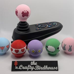 May include: A black joystick controller with a pink ball-shaped topper featuring a butterfly design. Five decorative balls in various colours, including blue with tulips, red with hearts, and green with a sheep design. The Crafty Birdhouse logo is at the bottom.