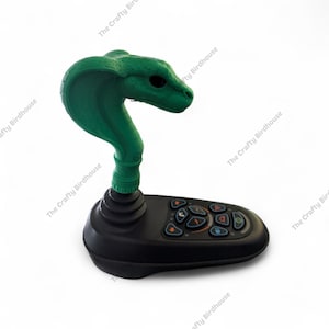 May include: A green cobra-shaped joystick with a black base. The cobra head is raised, and the base features several coloured buttons. The Crafty Birdhouse is printed on the side.