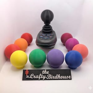 May include: A black joystick controller with a spherical top, surrounded by ten colourful spherical knobs in red, orange, yellow, green, purple, blue, pink, and magenta. A black rectangular sign in front displays the text the Crafty Birdhouse.