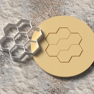 May include: A white metal honeycomb cookie cutter next to a tan, round, honeycomb-shaped stamp. The objects are on a light gray and brown textured surface, suggesting a baking or craft theme.