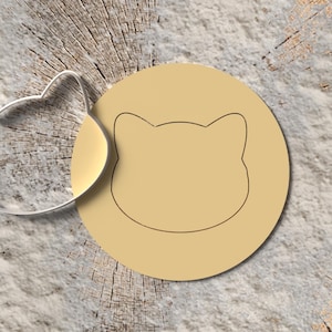 Könnte beinhalten: Ein silberfarbener Ausstecher in Katzenform neben einem beigefarbenen, kreisförmigen Keks mit einer Katzenumrisszeichnung. Die Objekte befinden sich auf einer strukturierten, hellbraunen Oberfläche, möglicherweise Holz.