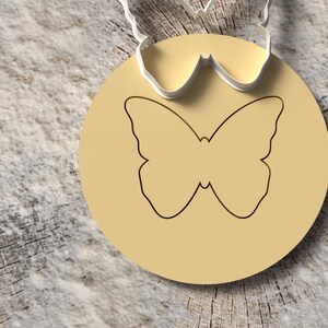 May include: A round, light tan pendant with a butterfly outline. The pendant is attached to a silver-colored metal piece with a butterfly shape. The background is a textured, light brown surface.