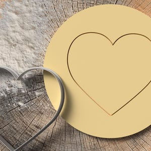 Könnte beinhalten: Ein herzförmiger Ausstecher neben einem herzförmigen Keks auf einer Holzoberfläche. Der Keks ist hellgelb. Mehl ist auf das Holz gestreut. Der Keks ist ein Kreis mit einem ausgeschnittenen Herzen.