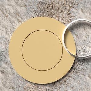 Puede incluir: Un plato beige con un borde circular negro y un anillo plateado. El plato está sobre una superficie texturizada y clara con un patrón de vetas de madera.