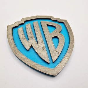 Puede incluir: Un logotipo de Warner Bros. impreso en 3D con forma de escudo. El escudo es plateado con un centro azul y las letras "WB" son en relieve y texturizadas.
