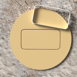 May include: A light tan, circular cookie cutter with a rectangular cutout in the center. A rectangular cookie cutter with a white border is placed on top. The background is a wood grain.