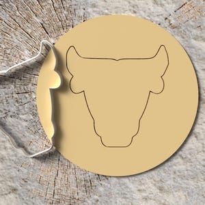 Könnte beinhalten: Ein klarer Ausstecher in Form eines Bullenkopfes neben einem beigefarbenen Kreis mit einer Bullenkopfkontur. Die Artikel befinden sich auf einer hellbraunen, strukturierten Holzoberfläche.