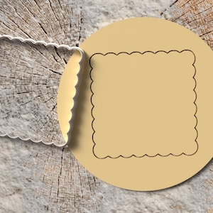Könnte beinhalten: Ein Ausstecher und ein Keksstempel werden auf einer Holzoberfläche gezeigt. Der silberne Ausstecher hat eine gezackte quadratische Form. Der beige Keksstempel hat ein passendes gezacktes quadratisches Design.